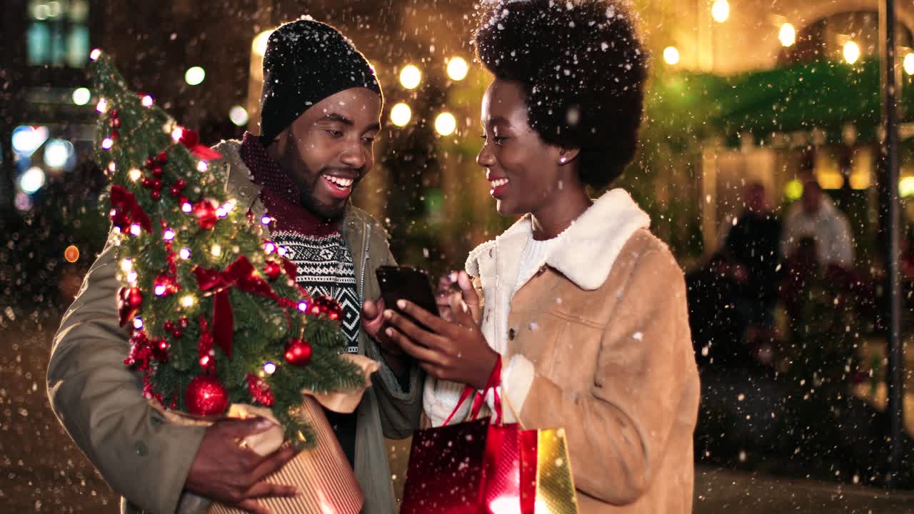 Close-up view of joyful African American couple talking and watching something on the phone while it¬¥s snowing on the street in Christmas