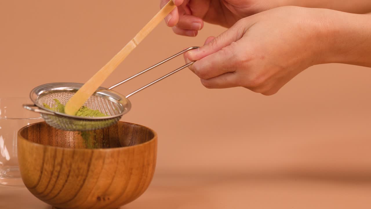 Hands sift matcha powder with bamboo scoop and strainer, minimal Japanese tea ceremony setup