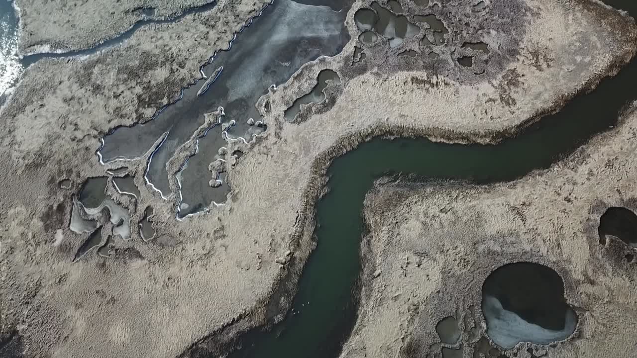 Overhead drone view of a sinuous tidal creek winding through winter salt marsh, with salt pannes, tidal pools and thin ice along the edges, Great Marsh–Essex Bay, MA