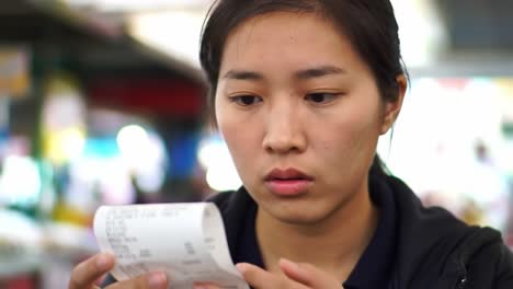 a young woman analyzes a receipt with concern, reflecting on her spending in a bustling marketplace setting, highlighting financial awareness and budgeting.