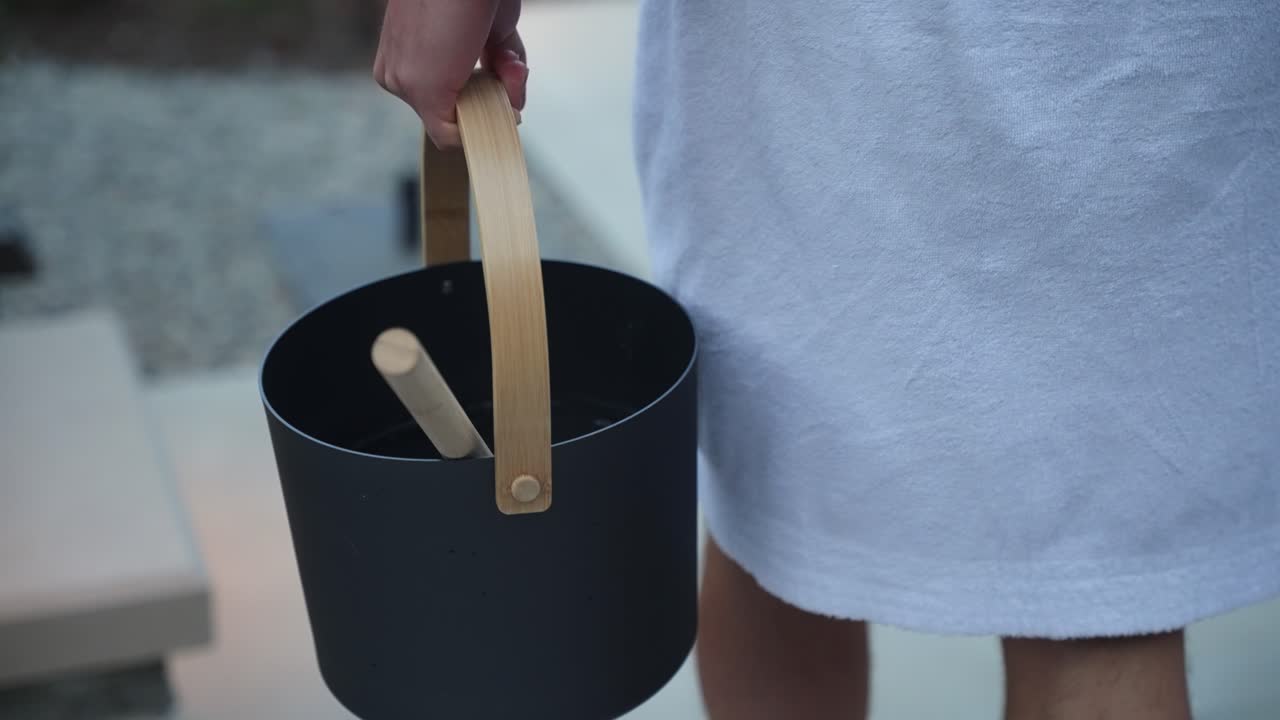 Close up of a white man dressed in a white dressing robe carrying a premium metal sauna water bucket with a wooden spoon walking down stairs at dusk