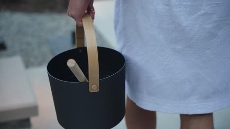 close up of a white man dressed in a white dressing robe carrying a premium metal sauna water bucket with a wooden spoon walking down stairs at dusk