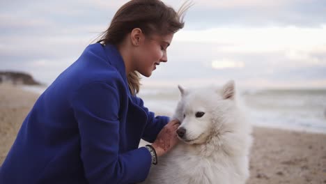 Junge-Frau-Sitzt-Im-Sand-Und-Streichelt-Ihren-Hund-Der-Rasse-Samojede-Am-Meer.-Weißes,-Flauschiges-Haustier-Am-Strand,-Das-Spaß-Hat