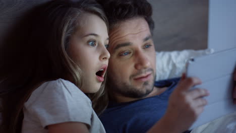 close-up view of little cute girl lying on the bed before sleeping with her handsome father and watching scary video on the tablet