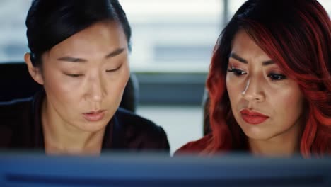 Two women sit closely at a desk, intensely focused on a computer screen in a modern office setting during late afternoon. Their expressions show concentration and determination.