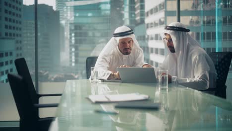 dos empresarios emiratíes exitosos en kandura tradicional blanca sentados en una reunión de oficina, negociando y hablando sobre oportunidades financieras, usando una computadora portátil.