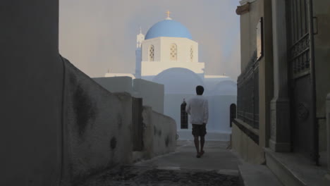 a man walks towards a greek orthodox cycladic style church with a blue dome