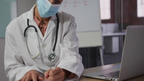 doctora caucásica con máscara sentada en el escritorio en la sala de reuniones tomando notas