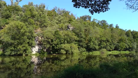 Tranquilo-Río-Sazava-Desde-Su-Orilla-Con-Una-Roca-Y-Un-Bosque-Al-Fondo