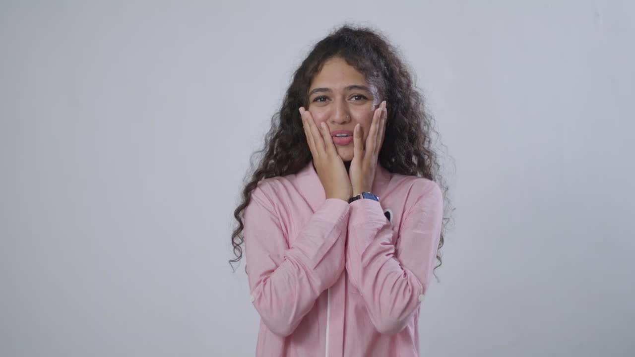 A South Asian woman with curly brown hair is happily surprised, with her hands pressed against her cheeks