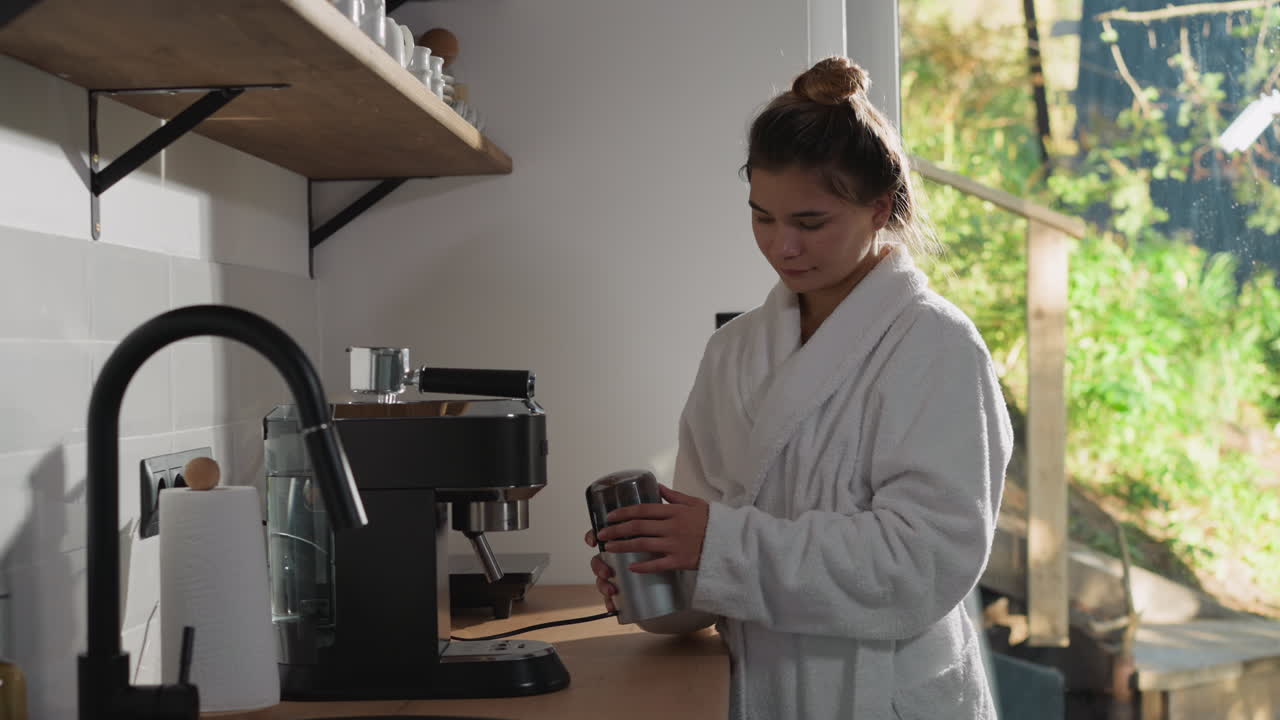 una joven mola granos de café en la cocina. una mujer con bata de baño suave hace polvo de espresso usando un molinillo. preparación de una bebida energética aromática antes del trabajo