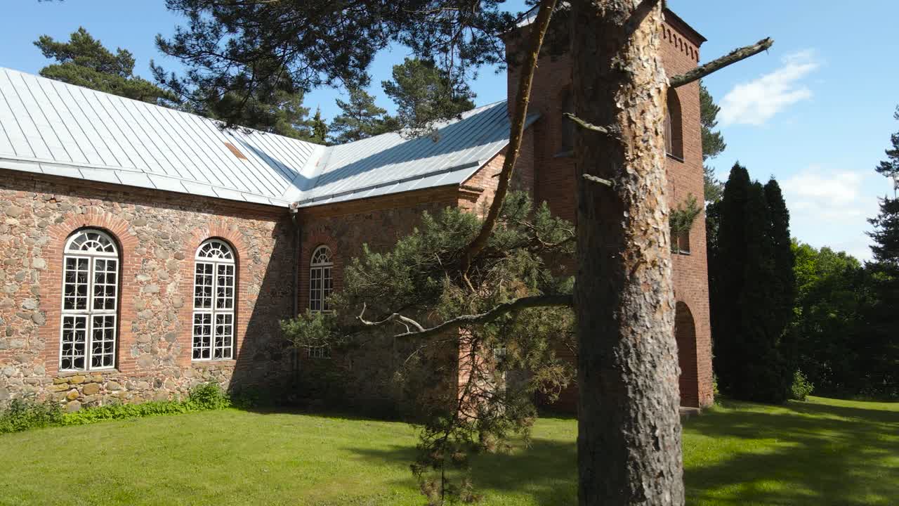 Aerial drone rotating around the Alatskivi church revealing beautiful rustic patterned stone walls, stonework, arched windows and scenic entrance. Surrounded by vibrant greenery and tall pine trees
