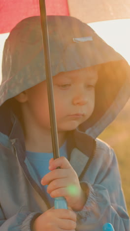 sad boy clutches umbrella tightly in field. small child expression mirrors heaviness of rainfall on riverbank. child somber mood in dreary surroundings