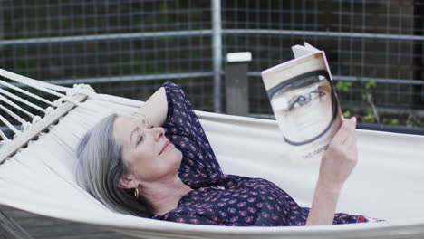 Mujer-Caucásica-De-Mediana-Edad-Leyendo-Un-Libro-Relajándose-En-Una-Hamaca-En-La-Terraza-En-La-Naturaleza,-Cámara-Lenta