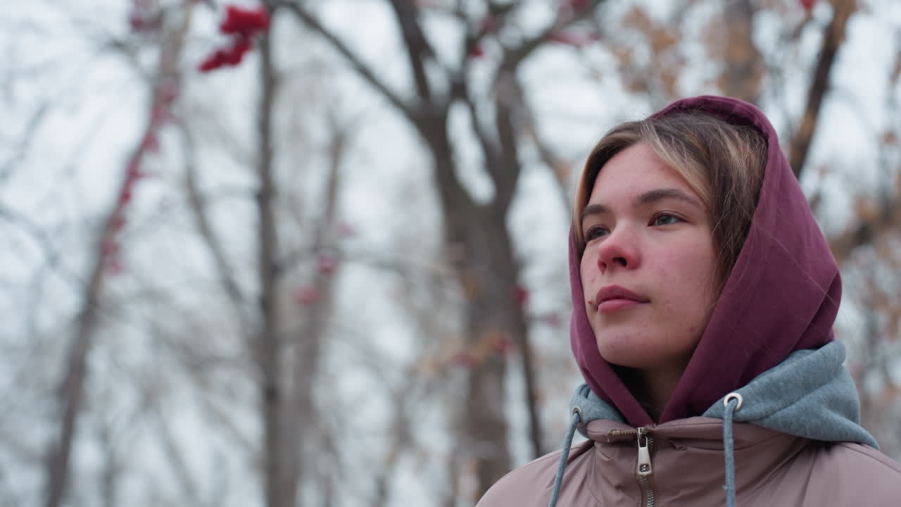 primer plano de una mujer elegante con una chaqueta de invierno con capucha caminando contemplativamente, mirando pensativamente a su entorno, árboles desnudos con rastros de nieve y luz suave