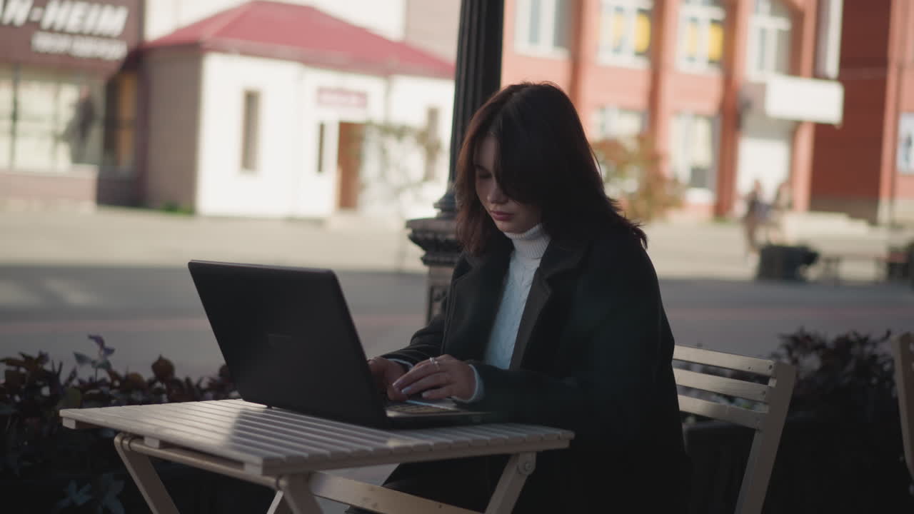 Woman working on laptop outdoors, hair cascading over her shoulders, set against blurred red brick building and greenery with distant figure moving in background