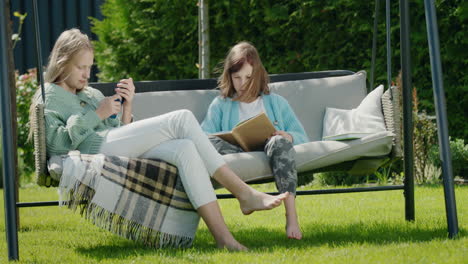 two friends are relaxing on a garden swing. one girl uses a smartphone, another reads a book