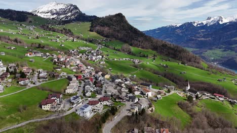 Drone-shot-showing-idyllic-village-on-green-mountain-and-snowy-alps-with-lake-in-background