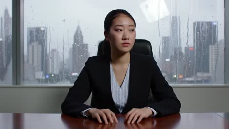 a professional woman in formal attire sits quietly at a desk, looking thoughtfully towards rain-soaked city skyscrapers. the atmosphere reflects a moment of contemplation amid a busy urban backdrop.