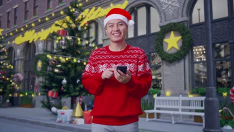 asian man in santa suit using smartphone and smile to camera while standing in christmas night