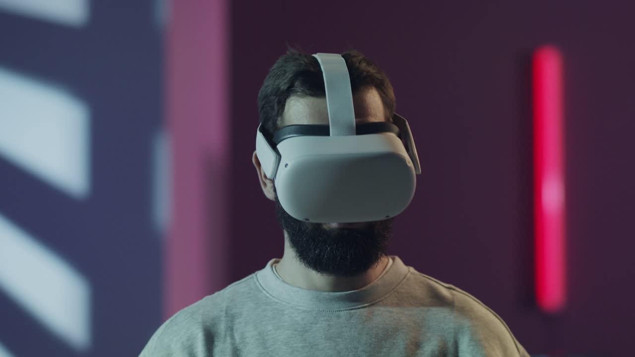 A bearded man wearing a VR headset looks around, fully immersed in a virtual experience. The futuristic room with neon lights enhances the theme of the metaverse and modern technology.