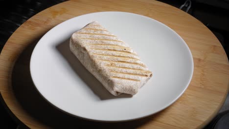 A-close-up-shot-of-a-plate-on-a-wooden-kitchen-table,-as-a-chef-carefully-places-a-healthy-toasted-tortilla-wrap-onto-the-plate-using-his-hand-and-a-spatula