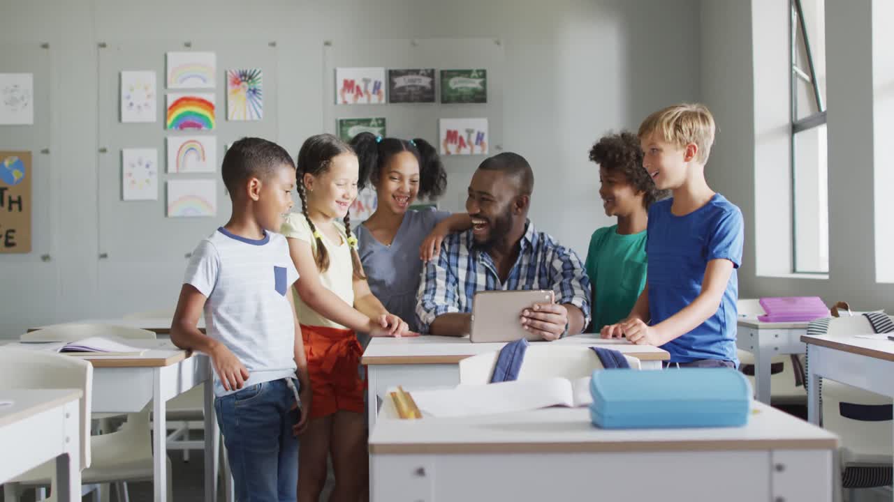 video de un feliz maestro afroamericano y una clase de alumnos diversos trabajando en una tableta