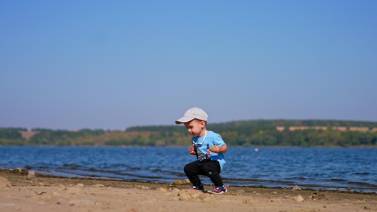 Smiling Caucasian baby boy in cap walks by the beach on sunny day. Toddler picks a stone and goes to throw it to the water.