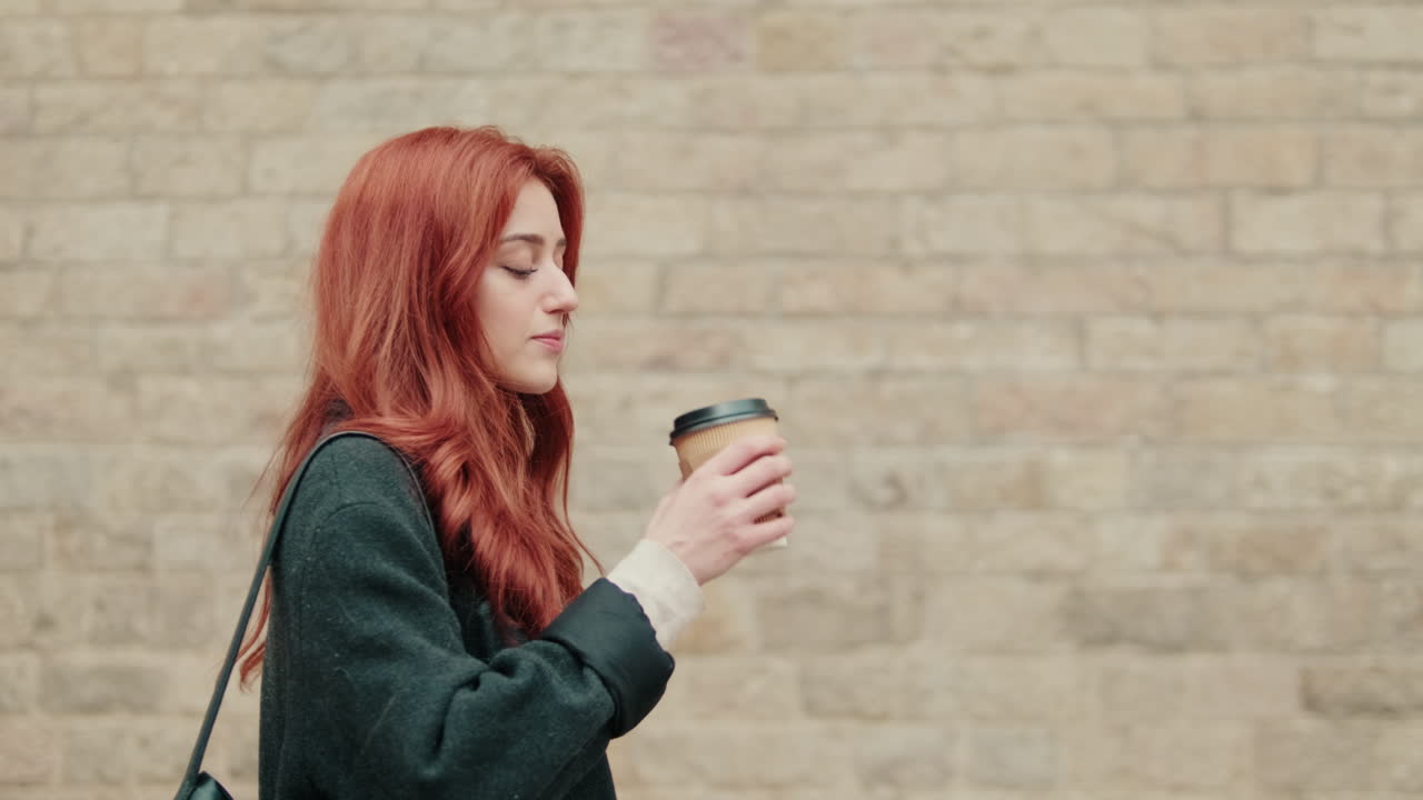 Giovane donna dai capelli rossi che cammina e beve caffè accanto a un muro di mattoni