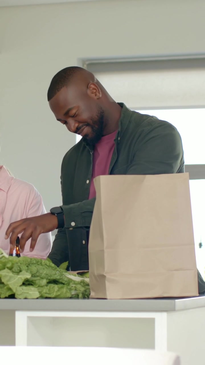 Vertical video: Grabbing bottle, man in green shirt sorting food on kitchen island, bag and lettuce