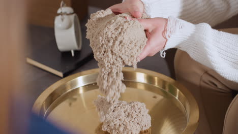 high angle of inner explorer seated on sofa gently smoothing soft wet soil mold with both hands during calming therapy session, showcasing mindful tactile interaction on golden tray
