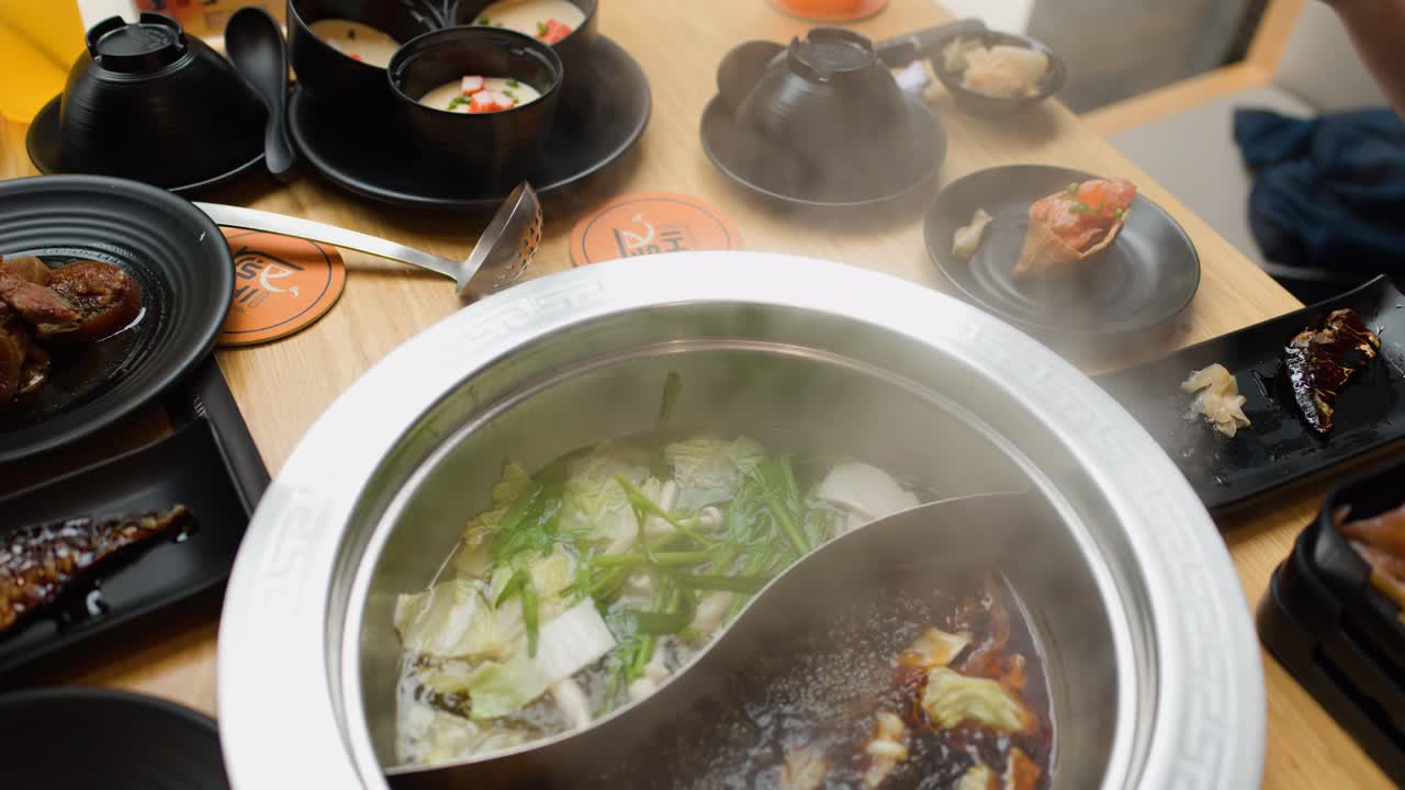 Hands add sliced meat to steaming dual broth hotpot surrounded by vegetables, sauces, drinks