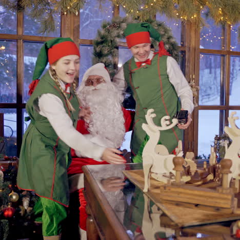 happy elves with wooden toys. many handmade toys on the table. santa claus having joyful time with fairy characters in the christmas decorated room.
