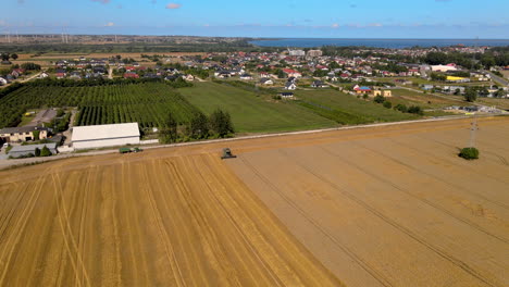 drone approaching an agricultural harvesting machinery at work on the wheat field in puck, poland - aerial tilt down