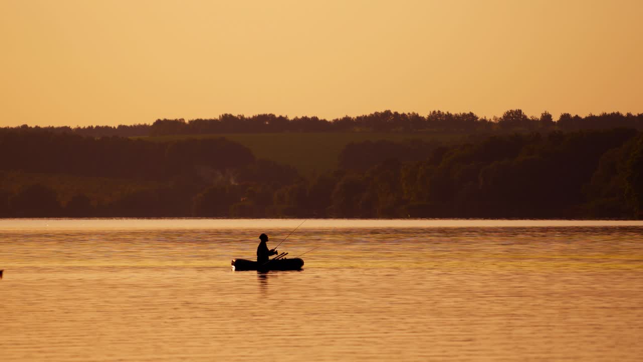 Silhouette of a man with fishing rods in the middle of the lake at sunset. Natural river background in the evening and a fisherman in a boat.