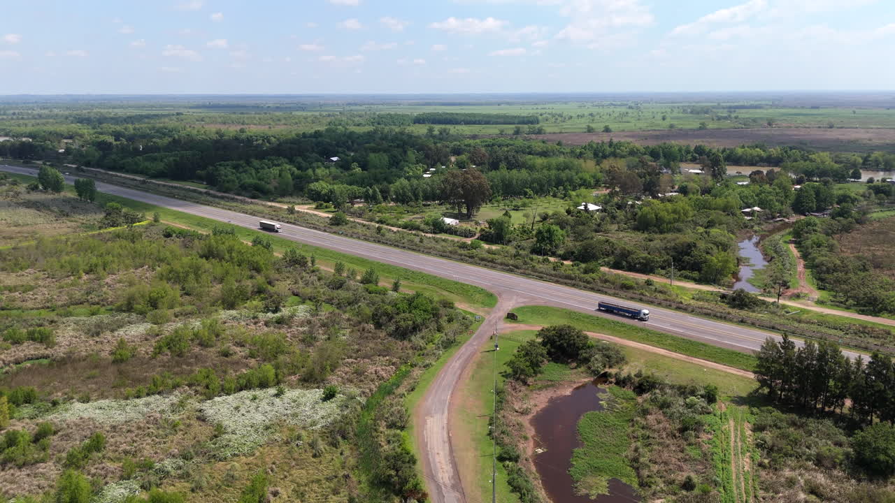 Aerial view capturing trucks driving along a highway, traversing the picturesque countryside of Misiones, Argentina, alongside a river and vibrant, lush forests on a sunny day
