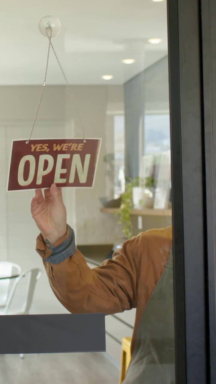 Vertical video: Opening shop, woman wearing green apron placing sign on glass door, copy space