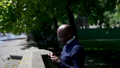 Black-male-standing-beside-a-wall-next-to-a-riverside,-using-a-smartphone