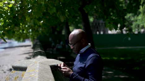 hombre negro de pie junto a una pared al lado de una orilla del río, usando un teléfono inteligente