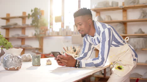 Happy-african-american-man-with-beard-using-smartphone-and-smiling-in-pottery-studio,-slow-motion