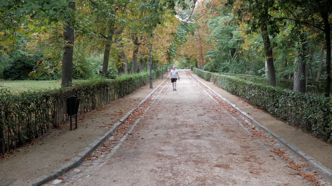 Senior male walking along path in Autumn surrounded by green and orange foliage in park. Health and minfulness habits on retirement for longevity.