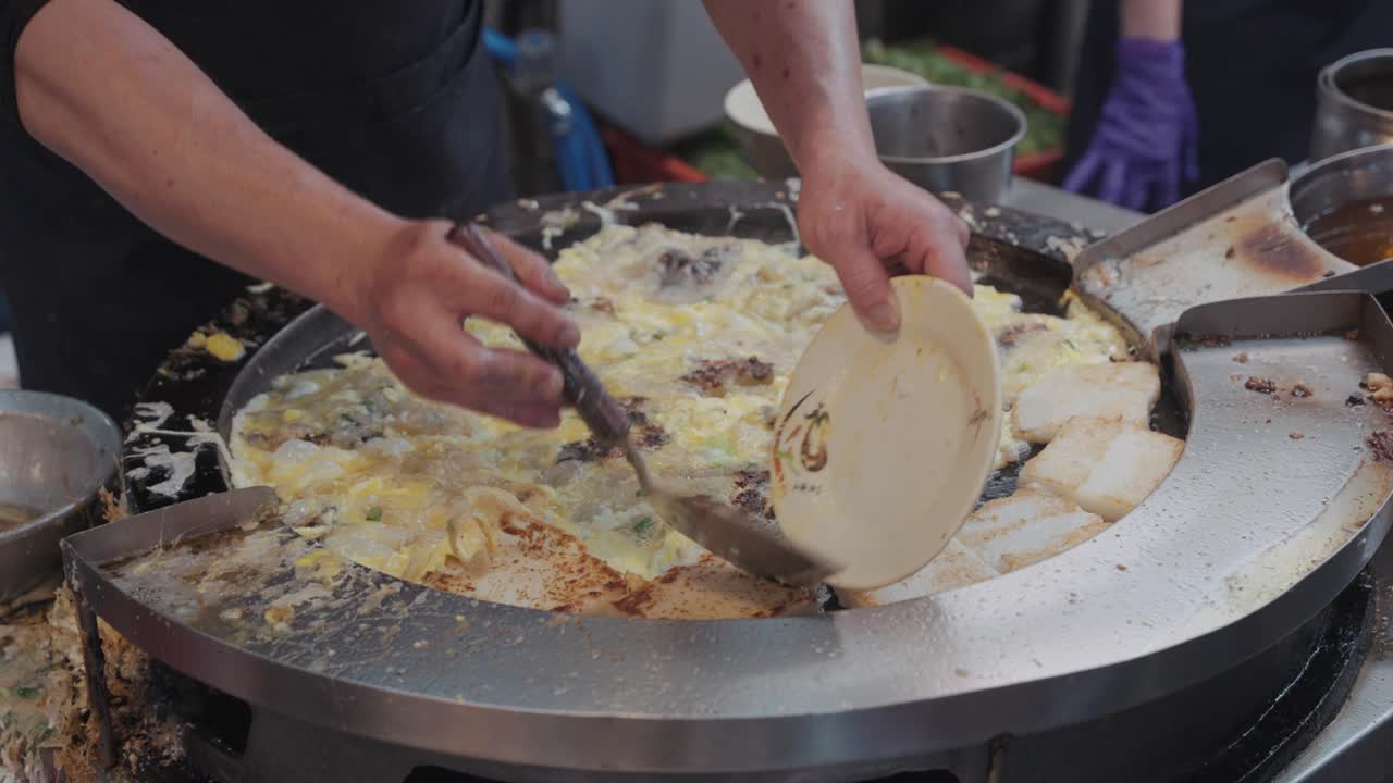Vendor frying and flipping sizzling Taiwanese oyster omelette on a hot round griddle, with eggs, oysters, and batter bubbling together into a savory street food delicacy