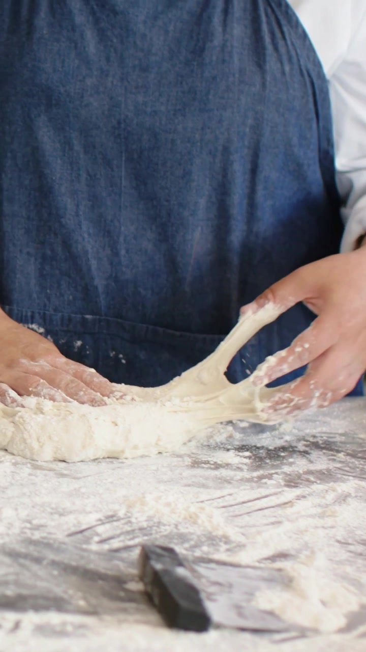 Video verticale: Fornaio con grembiule che sparge farina, piega e impasta l'impasto con una spatola in panetteria per fare il pane.