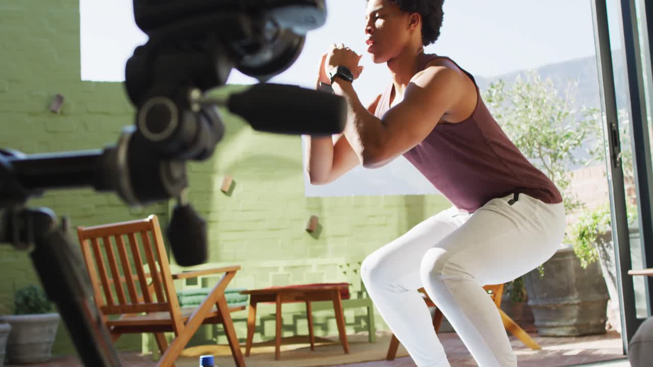 Uomo afroamericano atletico che si allena a casa, facendo squat e filmando un vlog di fitness.