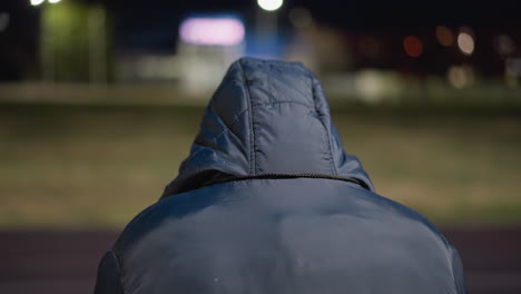 close-up back view of a man wearing a hood, seated alone in a quiet setting, the background is blurred and dimly lit