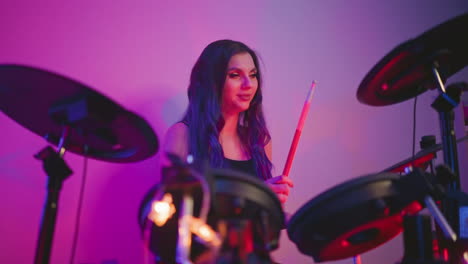 woman with long hair playing electronic drums in music studio surrounded by soft purple pink lighting, holding drumstick with neutral expression during indoor session, drum pads and cables in view