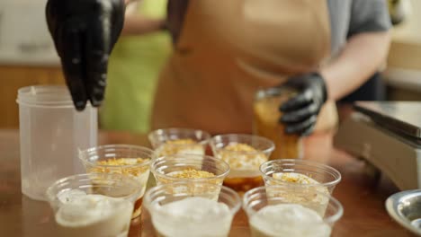 a person in black gloves is preparing a layered dessert with whipped cream and toppings.