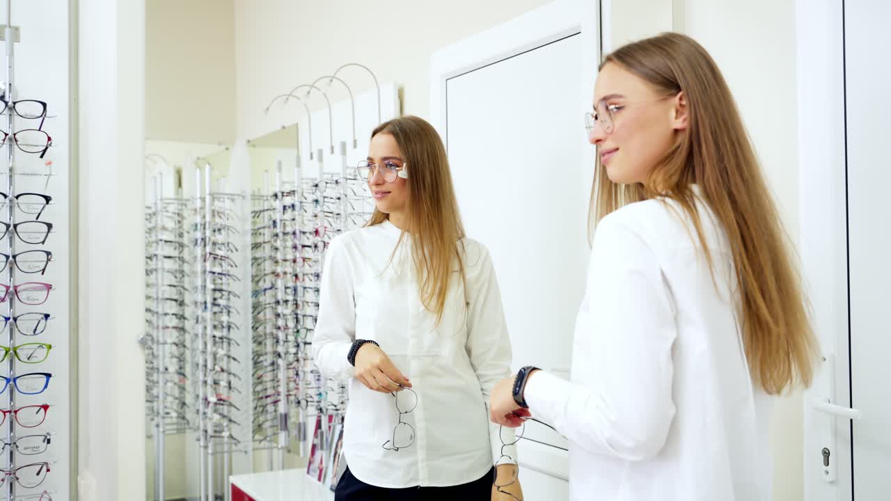 Girl at the optical store. Pretty woman in new spectacles looks at her reflection in the mirror. Eyeglasses choice.