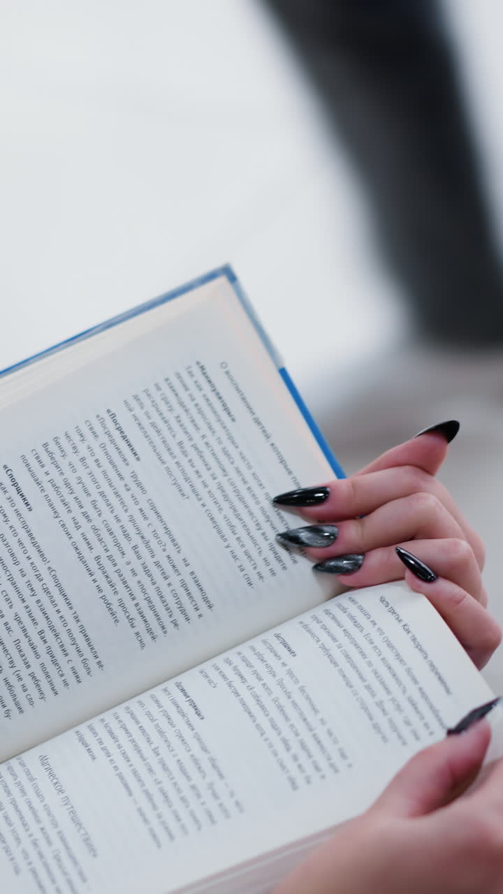 Close-up partial view of woman reading book while seated on bench, elegant black nails contrast with crisp pages, bright reflective lights create a soft ambiance