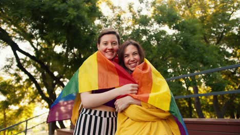 una chica con un corte de pelo corto en un pantano púrpura y una chica morena en un suéter amarillo amarillo se envuelven bajo la bandera de la comunidad lgbt